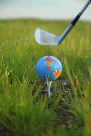 A miniature globe on a tee at a golf course. A golf club is poised behind the globe ready to swing.の写真素材