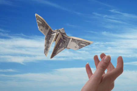 A hand trying to catch a dollar bill shaped like a butterfly. A blue sky serves as the background.の写真素材