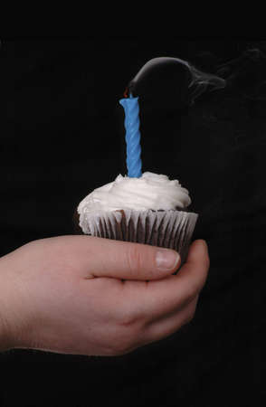 A hand holding a cupcake with one blue candle in it. The candle has just been blown out. Black background.の写真素材