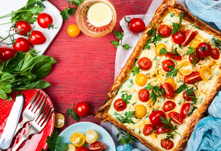 Deliciously simple tomato tart made with puff pastry, red and yellow cherry tomatoes, spinach and ricotta cheese. The perfect appetizer for summer. Concept of healthy eating or vegetarian food on red rustic wooden background, top view.の写真素材