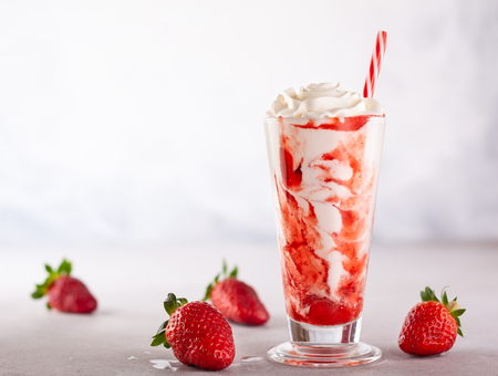 Strawberry milkshake with whipped cream and berry syrup in the tall glass on light background.