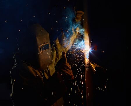 welder worker welding metal by electrode with bright electric arc and sparks during manufacture of...
