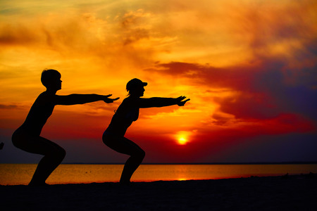 Yoga people training and meditating in warrior pose outside by beach at sunrise or sunset. Woman and man yoga exercising training in serene ocean landscape. Silhouette of couple against sun.の写真素材