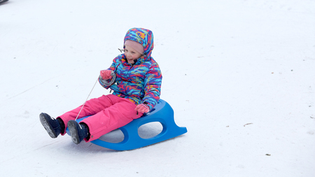 Cheerful girl riding a sled downhill on a snow covered sledge trail in a white sunny winter mountain landscapeの写真素材
