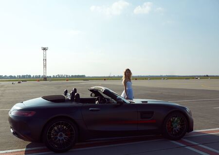 The elegant blonde beautiful woman posing near luxury vehicle on planes background. Girl wearing...