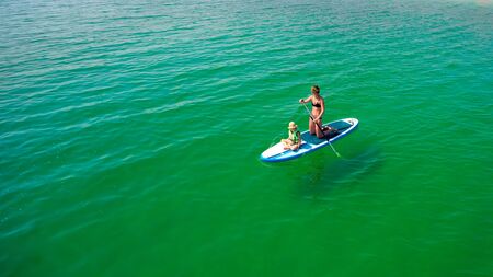 Little girl in a life vest sitting on the paddle board with mother.の写真素材