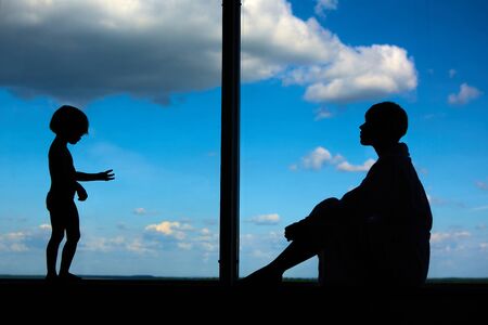 Silhouettes of Mom with a daughter of 3 years in front of a large window with blue sky and clouds. Stay at home for the quarantine prevention of the coronavirus pandemic. Self-isolationの写真素材