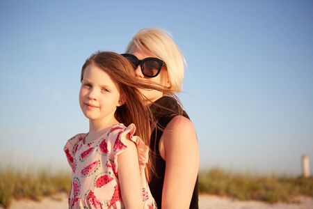 Smiling mother and beautiful daughter having fun on the beach. Portrait of happy woman giving a piggyback ride to cute little girl with copy space. Portrait of kid embracing her mom during summer.の写真素材