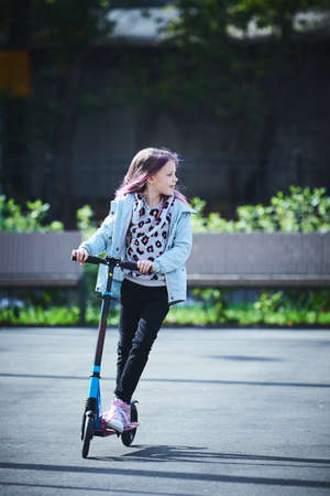 Happy beautiful girl is riding along the street on the kick scooter on a windy day. Baby having free time playing. The concept of a healthy lifestyle. Kids sportの写真素材