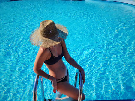 Portrait of a beautiful woman getting out of a swimming pool. beautiful blond hair tanned female model posing by blue pool water. Outdoor summer portrait of sexy girl in straw hat an sunglasses.の写真素材