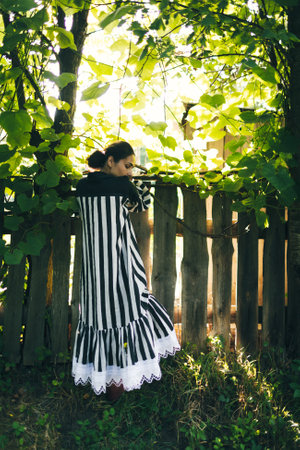 Stylish young indian woman in black and white dress on a background of green trees and aged wooden fence. Boho woman relaxing in countryside, simple slow life style. Atmospheric imageの写真素材