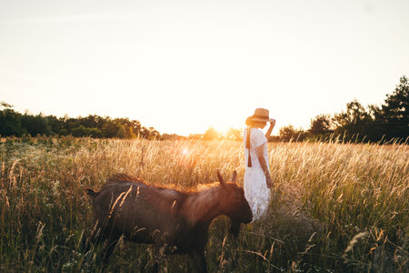 Summer delight and travel. Young carefree female in rustic linen cloth relaxing in summer meadow. Stylish boho woman with straw hat posing among wildflowers in sunset light. Atmospheric moment.の写真素材