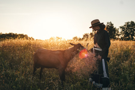 Young woman in the field is grazing her white goats. The girl and goats in the meadow in summer. Love for animals. Goat farm. Pets. Happy woman with animal. Kindness and love for animals. Kisses a petの写真素材