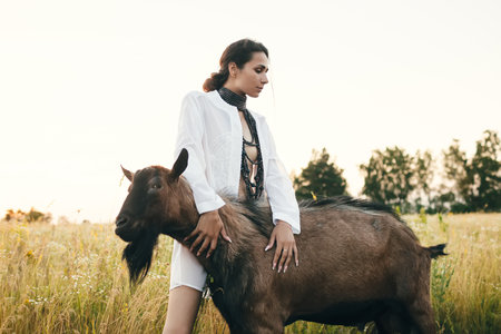 Young woman in the field is walking with goats. The girl and goats in the meadow in summer. Love for animals. Goat farm. Pets. Happy woman with animal. Kindness and love for animals. Kisses a petの写真素材