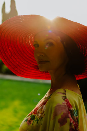 Portrait of a woman wearing a large red sun hat during golden hour. The soft, warm sunlight highlights the edges of the hat and casts a beautiful glow on her face, creating a serene and tranquil atmosphereの写真素材