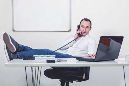 Relaxed businessman with legs on the table talking and looking at computerの写真素材