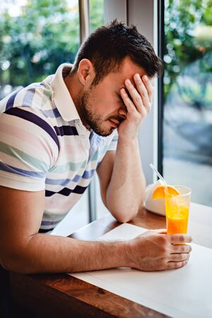Man with hangover  drinking orange juice in a cafe and holding headの写真素材
