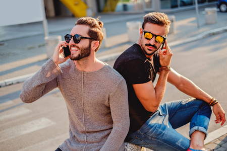 Two friends sitting in the street, talking on the phone. One is happy and one is sadの写真素材