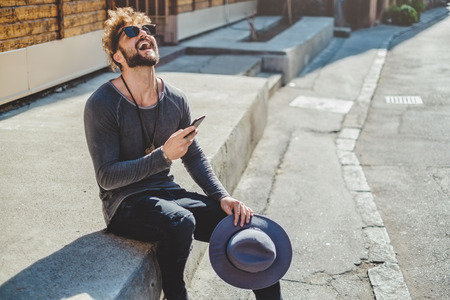 Hipster laughing while holding mobile phone and sitting on the stairsの写真素材