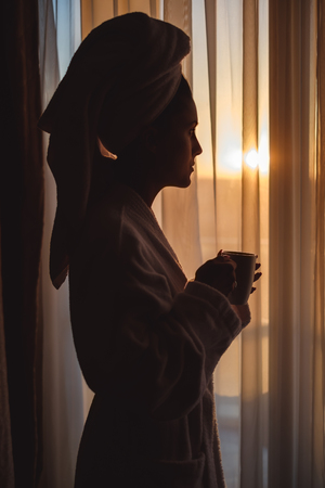 Woman in bathrobe looking through window into a sunset and holding cupの写真素材