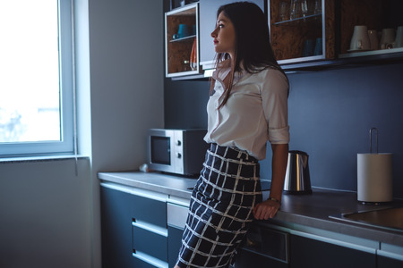 Sad girl leaning on a kitchen countertop looking through the windowの写真素材