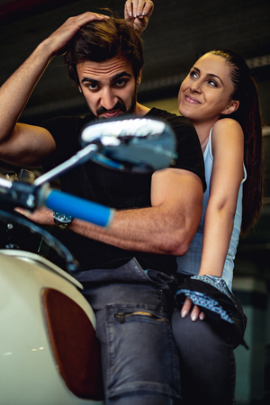 Biker looking into a mirror of his bike while smiling girlfriend is messing up his hairの写真素材