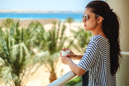 Beautiful girl enjoying cup of coffee on a balcony with sea view in the background
