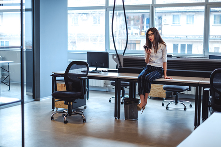 Angry girl looking at the phone while sitting on the desk in the officeの写真素材