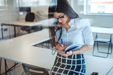 Sexy  businesswoman behind the glass writing in notebook  while sitting on the office deskの写真素材