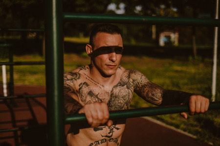 Tough tattooed man leaning on the workout bar outside and looking far awayの写真素材