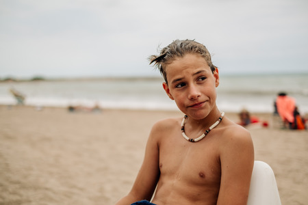 Wet serious boy sitting on the beach and thinkingの写真素材