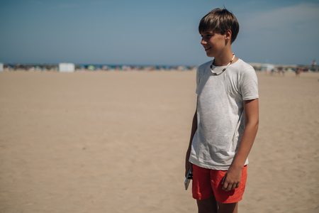 Boy standing on the sunny beach and smiling while looking far awayの写真素材