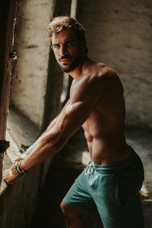 Athletic, bearded, shirtless man standing by the window with serious expressionの写真素材