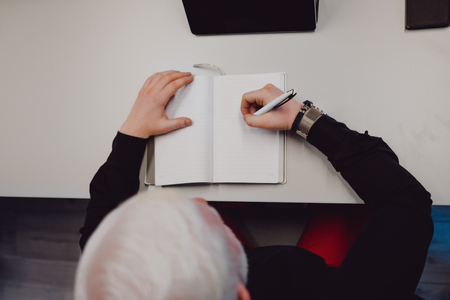 Older man with white hair at  the office desk writing in his notebookの写真素材