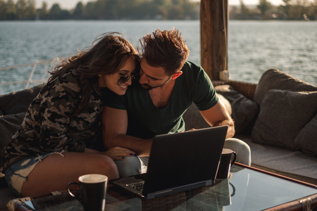 Happy couple smiling and working on laptop bu the river on a sunny dayの写真素材