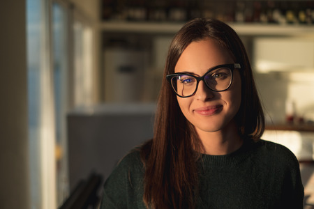 Beautiful brunette girl with glasses smiling and standing by the window on a sunny dayの写真素材