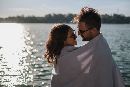 Smiling couple look at each other while covered with blanket by the riverの写真素材