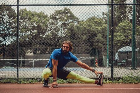 Bearded athlete man stretching leg in front of tennis court
