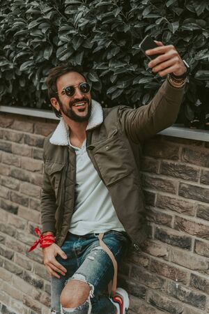 Happy, stylish, bearded man taking a selfie while leaning on the wall
