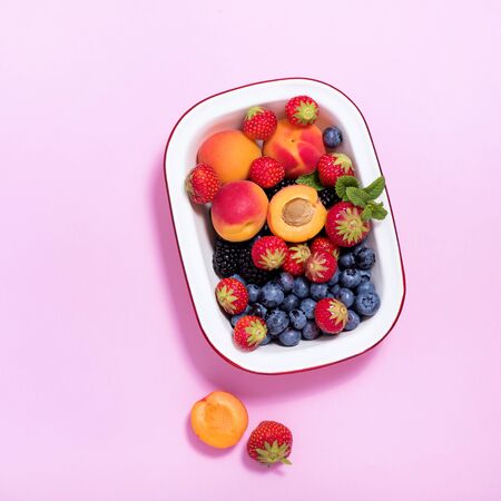 Ripe apricots and summer berries in a bowl on pink paper background, summer fresh fruits, vitamins, top view, vegan ingredientsの写真素材