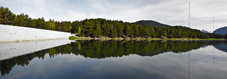 The Panorama of Engolasters lake in Andorra Pyrenees mountains. The dam is left at the fir forest and the sky Reflect in water and fthe view at mountain snowy peaks at right. .の写真素材