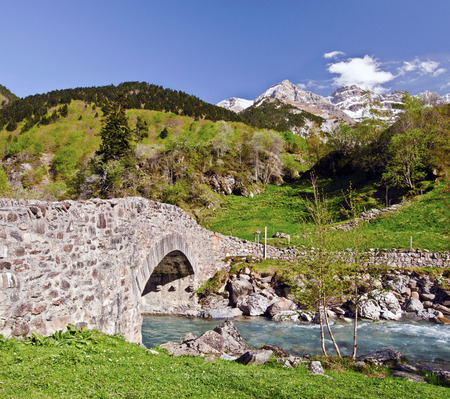 Nadau bridge through river Gave de Gavarnie is a stone bridge on the way from the village to the mountain Gavarnie Circus Ambama forest slopes and tops of snowy Pyrenees mountains at backgroundの写真素材