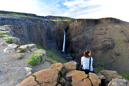 Young woman is sitting at the rock in front of the city of Litlanesfoss waterfall surrounded by vertical basalt columns, Fljotsdalshreppur municipality of Eastern Icelandの写真素材