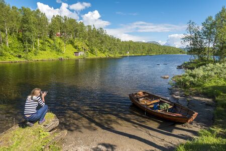 Young woman is photographing the lake and forest landscape in Norwegian Nordland. Fisher's boat in...