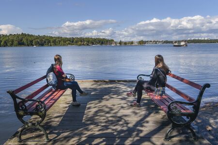 Stockholm, Sweden - August 18, 2017 Young women sitting in benches in the coast of Djurgarden island of Stockholm city facing to Isbladsviken bay.のeditorial素材