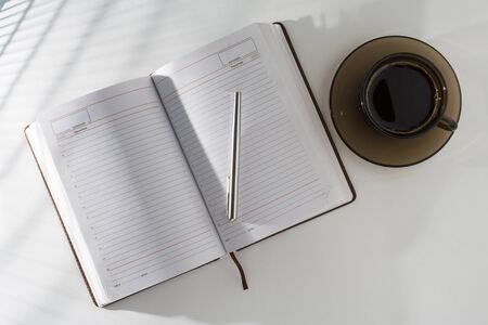 The table at the window, the light on the table through the blinds or lattice. On the table in an open diary and pen, standing next to a cup of coffee.の写真素材