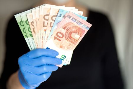 Senior woman wearing mask holds cash Euro notes in a hand in gloves. Selective focus.の写真素材