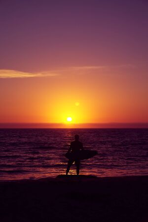 Surfer with surfing board on a seaside with beautiful sunset.