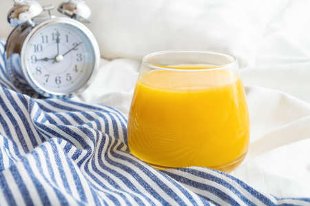 Glass of orange juice and alarm clock on bed with white linen and blue bedspread. Good morning concept.の写真素材