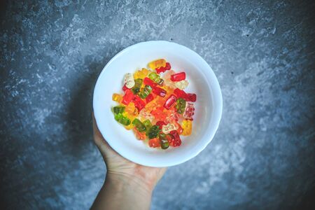 Colorful gummy bears in the white bowl holding in hand on the gray and white background with black vingete.の写真素材